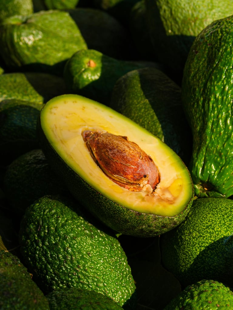 A halved avocado with its brown pit exposed, surrounded by whole green avocados in close-up view