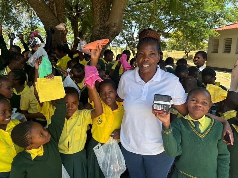 A woman in a white shirt smiles surrounded by school children in yellow and green uniforms outdoors under a large tree